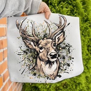 May include: A light gray tote bag featuring a detailed illustration of a deer's head with large antlers. The deer is depicted in shades of brown and tan, set against a backdrop of green and black paint splatters. The bag is held by a person.