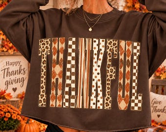 May include: A dark brown long-sleeve cropped sweatshirt featuring the word THANKFUL printed in large block letters. Each letter displays a unique pattern including leopard print, brown argyle, checkered, and polka dot designs.