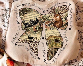 May include: A beige sweatshirt with a large bow design featuring a deer head, hunting scenes, and camouflage patterns. The text includes phrases like "Call of the hunt" and "Whisper of the woods."