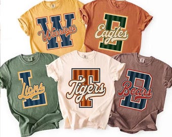 May include: Five short-sleeve crew neck t-shirts in yellow, orange, green, cream, and brown. Each shirt features a large varsity-style letter with vertical stripes and a cursive team name overlay: Warriors, Eagles, Lions, Tigers, and Bears.