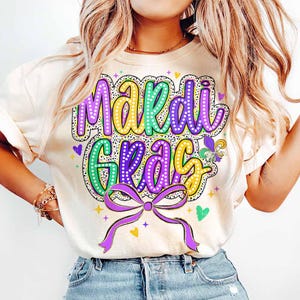 May include: A white t-shirt with a colourful "Mardi Gras" design. The design features purple, green and gold lettering with a sparkly effect. A purple bow with gold accents is below the lettering.