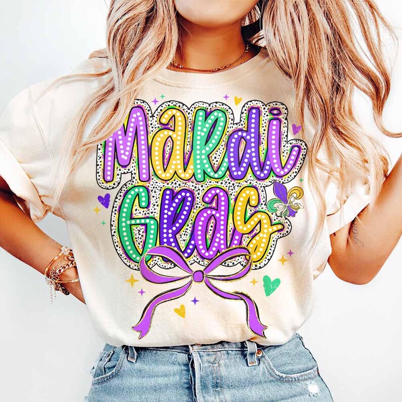 May include: A white t-shirt with a colourful "Mardi Gras" design. The design features purple, green and gold lettering with a sparkly effect. A purple bow with gold accents is below the lettering.
