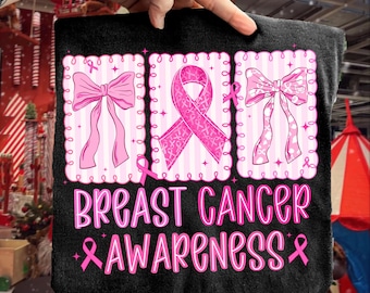 May include: Black t-shirt with a pink breast cancer awareness design. The design includes pink ribbons and bows, along with the words "BREAST CANCER AWARENESS" in a pink, stylized font. The shirt is held up by a hand.
