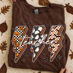 May include: A brown t-shirt featuring a Halloween-themed design. The design includes three lightning bolt shapes with the word "teacher" in the center. The lightning bolts are decorated with pumpkins, ghosts, and bats.