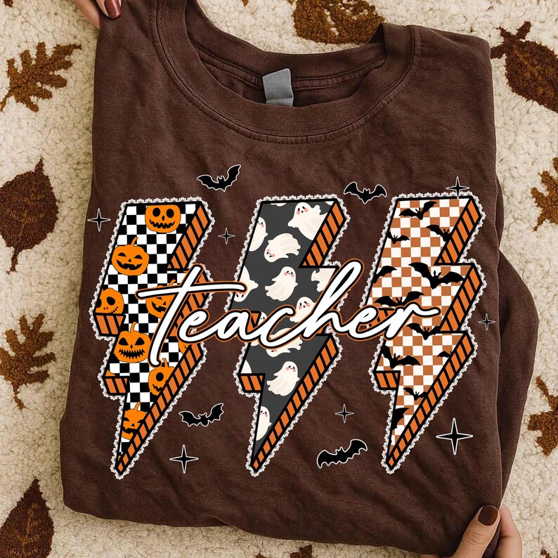 May include: A brown t-shirt featuring a Halloween-themed design. The design includes three lightning bolt shapes with the word "teacher" in the center. The lightning bolts are decorated with pumpkins, ghosts, and bats.