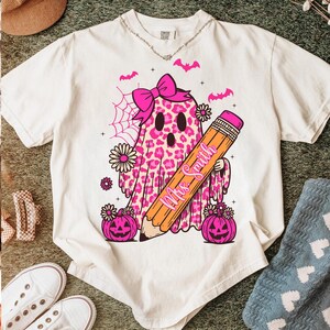 May include: Cream-colored t-shirt featuring a pink leopard print ghost with a bow, holding a pencil with "Mrs. Smith" on it. Pink pumpkins, flowers, and bats complete the Halloween-themed design.
