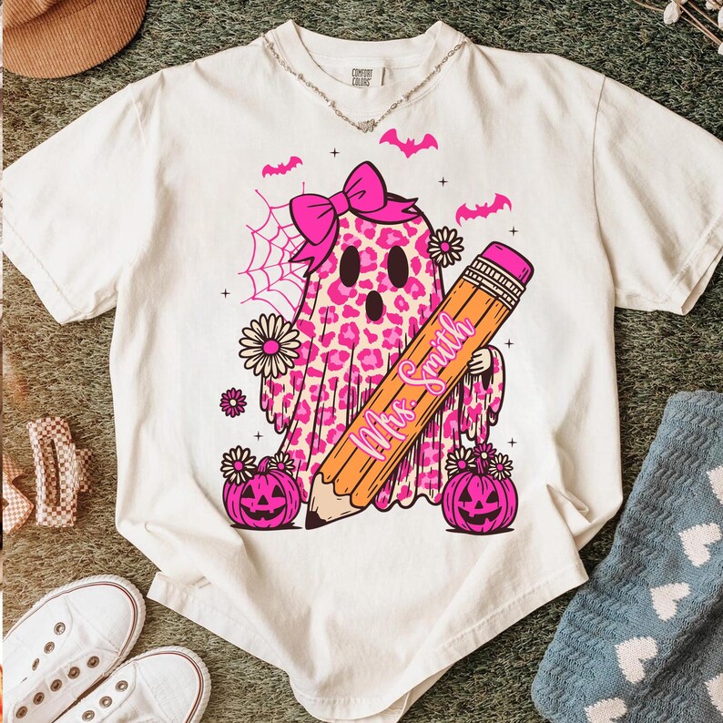 May include: Cream-colored t-shirt featuring a pink leopard print ghost with a bow, holding a pencil with "Mrs. Smith" on it. Pink pumpkins, flowers, and bats complete the Halloween-themed design.