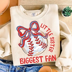 Op de afbeelding: Een beige sweatshirt met een rode en blauwe baseball-afbeelding en de tekst "Little Sister Biggest Fan".