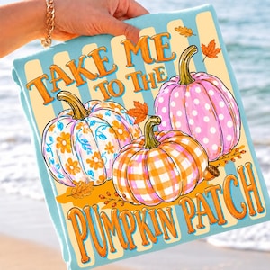 May include: A light blue blanket featuring three pumpkins with orange, pink, and white patterns including flowers, polka dots, and gingham checks. The text reads "TAKE ME TO THE PUMPKIN PATCH" in orange letters, accented by small orange autumn leaves.