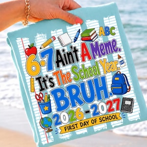May include: A light blue t-shirt featuring a graphic print of school supplies including a red apple, pencils, a notebook, a blue backpack, and a globe. The text reads 6-7 Ain't A Meme. It's The School Year. BRUH. 2026-2027 FIRST DAY OF SCHOOL.