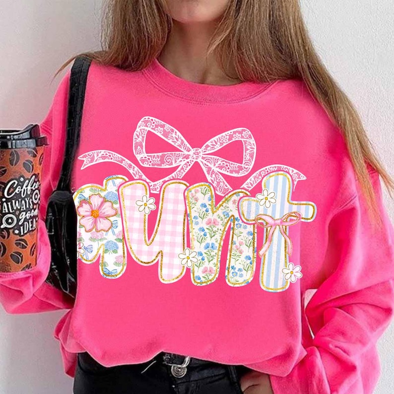 K&ouml;nnte beinhalten: Rosafarbener Pullover mit einem floralen Muster, auf dem "Tante" mit einer wei&szlig;en Schleife obenauf steht.