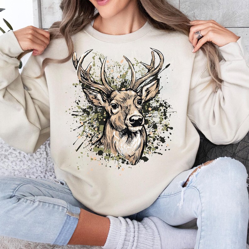 May include: A cream-colored sweatshirt featuring a detailed illustration of a deer's head with large antlers. The deer is set against a backdrop of green, black, and brown paint splatters. The sweatshirt is a casual, comfortable style.