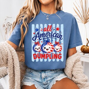 Op de afbeelding: Een blauw t-shirt met de woorden "all American DUMPLING" in rood en wit. Het shirt heeft een afbeelding van drie dumpling-personages met patriottische ontwerpen. De dumplings hebben lachende gezichten en zijn versierd met sterren en strepen.