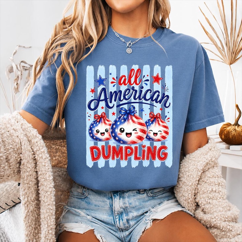 Op de afbeelding: Een blauw t-shirt met de woorden "all American DUMPLING" in rood en wit. Het shirt heeft een afbeelding van drie dumpling-personages met patriottische ontwerpen. De dumplings hebben lachende gezichten en zijn versierd met sterren en strepen.