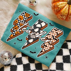 May include: Teal-colored Halloween-themed shirt with three lightning bolt designs. Each bolt features a different pattern: jack-o'-lanterns, ghosts, and bats. The word "teacher" is written across the bolts in a cursive font.