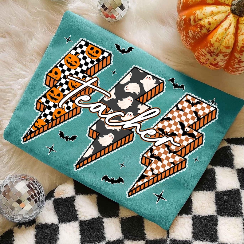 May include: Teal-colored Halloween-themed shirt with three lightning bolt designs. Each bolt features a different pattern: jack-o'-lanterns, ghosts, and bats. The word "teacher" is written across the bolts in a cursive font.
