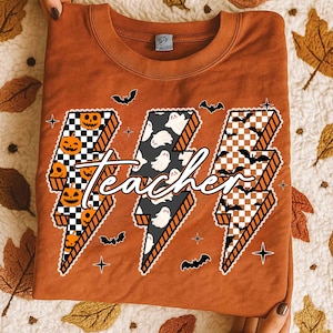 May include: An orange-brown sweatshirt featuring a Halloween-themed design. The design includes three lightning bolt shapes with checkered patterns, pumpkins, ghosts, and bats. The word "teacher" is written across the bolts in a cursive font.