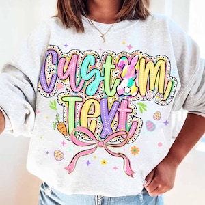 Op de afbeelding: Een lichtgrijs sweatshirt met een kleurrijk grafisch ontwerp met de tekst "Custom Text" met een konijn, eieren en een strik. Het ontwerp is omgeven door kleine sterren en stippen.
