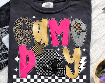 May include: Dark gray t-shirt with the words "GAME DAY" in a mix of leopard print, gold, pink, and silver glitter. A checkered football helmet is featured in the design, along with star and lightning bolt accents.