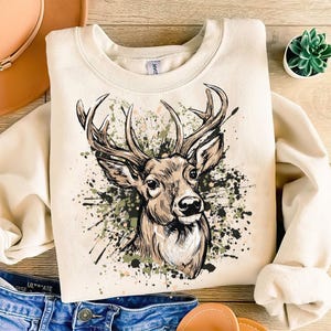 May include: Cream-colored sweatshirt featuring a detailed illustration of a deer's head with large antlers. The deer is set against a backdrop of green, black, and brown paint splatters. The sweatshirt is folded, with a glimpse of denim jeans and a brown leather bag.