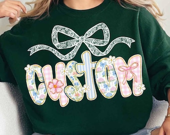 May include: A dark green sweatshirt with a white lace bow and the word "CUSTOM" in colorful patterned letters. The letters are decorated with flowers, gingham, and stripes.