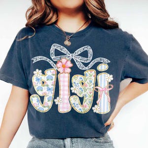 K&ouml;nnte beinhalten: Ein marineblaues T-Shirt mit einem floralen Design, das "Gigi" buchstabiert. Das Design enth&auml;lt eine wei&szlig;e Schleife mit Spitzenbesatz, rosa und blaues Gingham und kleine Blumen.