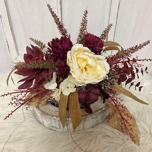 Könnte beinhalten: Blumenarrangement mit einer großen, cremeweißen Pfingstrose im Mittelpunkt, umgeben von tiefroten Chrysanthemen und anderen Blumen. Das Arrangement umfasst getrocknete Blätter und befindet sich in einer rustikalen, weiß getünchten Holzschale.
