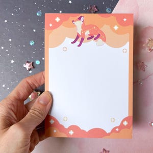 May include: A rectangular notepad featuring a white centre for writing, framed by a border of orange and pink clouds. At the top, a stylised orange fox is illustrated against the clouds, accented by small white four-pointed star icons.