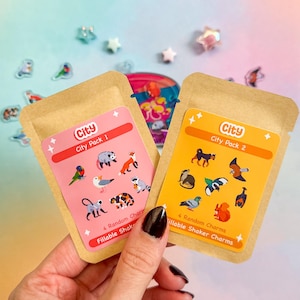 Könnte beinhalten: Zwei braune Papiertüten mit bunten Etiketten. Die Etiketten lauten "City City Pack 1" und "City City Pack 2". Jede Tüte enthält 4 füllbare Shaker-Anhänger mit Tierillustrationen.