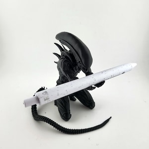 May include: A black Alien figurine holding a white pen with a speckled design. The figurine is in a kneeling position with its tail extended. The pen has a white body with a silver speckled design.
