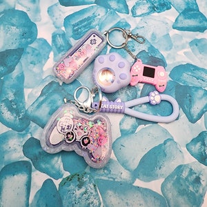 May include: A set of five keychains featuring a purple paw print, a pink and purple game controller, a clear keychain with a purple game controller and glitter, a clear keychain with a purple and pink game controller and glitter, and a clear keychain with a purple and pink game controller and glitter. The keychains are attached to a silver chain and a purple strap with the text "CAT STORY".