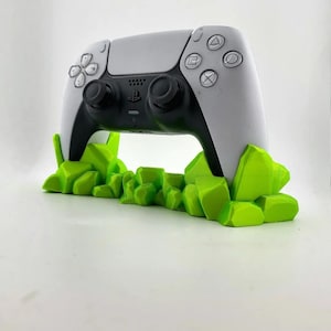 May include: A white and black game controller rests on a bright green, rocky-shaped stand. The controller has black joysticks and buttons with the PlayStation symbols. The stand is a vibrant, eye-catching accent for the gaming setup.