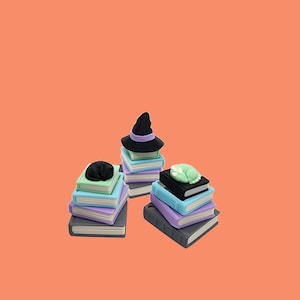 May include: Three stacks of miniature books in shades of gray, purple, blue, and green, topped with a black cat, a witch hat, and a green cat. The background is a solid orange color.