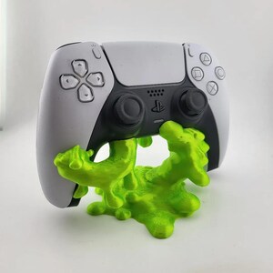 May include: A white and black video game controller resting on a bright lime green 3D-printed stand shaped like a puddle of liquid slime. The controller features standard directional buttons, two analog sticks, and the PlayStation logo in the center.