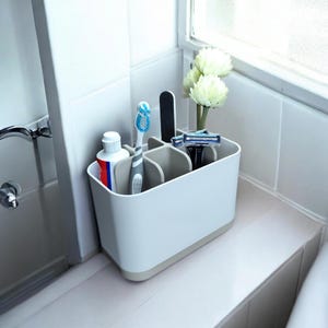 May include: A white and beige bathroom organizer with multiple compartments. It holds a tube of toothpaste, a toothbrush, a nail file, a razor, and a small bouquet of white flowers. The organizer is sitting on a white tiled surface.