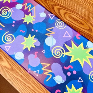 May include: A colorful desk mat with a space-themed design. The mat features a purple and blue background with various shapes, including stars, planets, and swirls. The design is accented with yellow, green, and pink elements. The mat is rectangular and rests on a wooden surface.