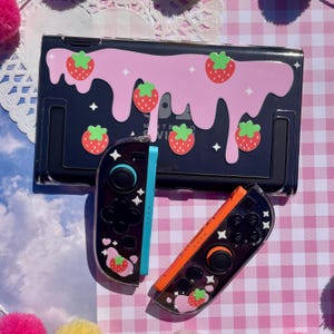 以下が含まれることがあります： イチゴをテーマにした黒いNintendo Switchケースとコントローラーのセット。ケースは、赤いイチゴと白い星が付いたピンク色のドリップデザインです。コントローラーは、イチゴと星のディテールが施された黒とオレンジ色です。