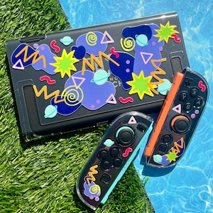 Peut inclure: Un étui noir pour Nintendo Switch et deux manettes Joy-Con décorés de formes abstraites colorées. L'étui présente des planètes violettes, des étoiles jaunes et des zigzags bleus. Une manette a un côté orange, l'autre un côté turquoise.
