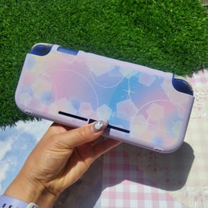 May include: A handheld gaming console protective case featuring a pastel gradient of light purple, pink, and blue. The surface is decorated with a pattern of white circular outlines, geometric shapes, and small starburst accents across the back.