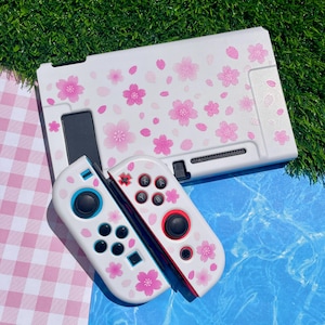 May include: A white gaming console dock and two matching controllers featuring a pink cherry blossom pattern. The controllers have black joysticks, buttons, and directional pads, with one controller featuring a red ring around the joystick.