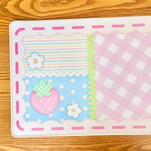 Peut inclure: Un tapis rectangulaire aux couleurs pastel avec un design fantaisiste. Il présente une fraise, des fleurs et un motif vichy. Le tapis a un bord festonné et une section bleue à pois.