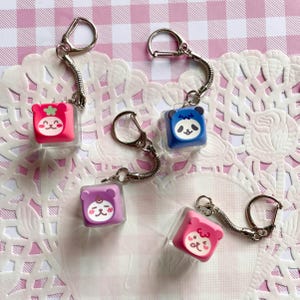 May include: Five colorful square keychains with cartoon animal faces. The keychains are pink, blue, and purple, with silver metal hardware. Each keychain has a different animal face design, including a panda, bear, and other cute characters.