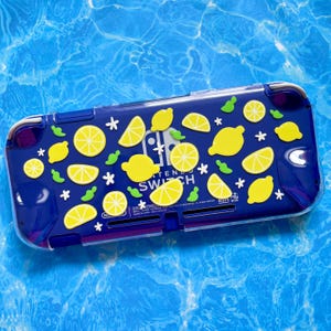 May include: A blue Nintendo Switch Lite console with a lemon-themed design. The case is decorated with yellow lemons, lemon slices, green leaves, and white star accents. The text "NINTENDO SWITCH" is visible.