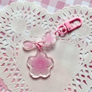 Puede incluir: Un llavero acrílico rosa con un diseño de flor de cerezo. El llavero tiene una apariencia clara y transparente con un diseño de flor rosa y un cierre rosa. El llavero está sobre un mantelito de encaje blanco.