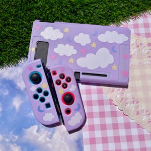 May include: A lavender gaming console and two matching controllers featuring a pattern of white clouds, yellow stars, blue crescent moons, and pink hearts. The console and controllers have a smooth finish with black buttons and joysticks.