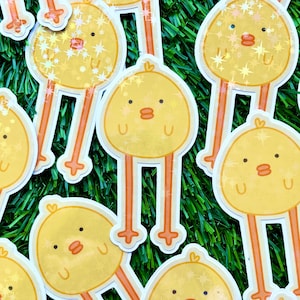 May include: A set of yellow chick stickers with glitter accents. The chicks have black eyes and beaks and are standing on two legs.