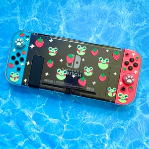 May include: A blue and black Nintendo Switch console with a custom design featuring green frogs, red strawberries, and white paw prints. The console has the text "Nintendo Switch" and "Nom" printed on it.