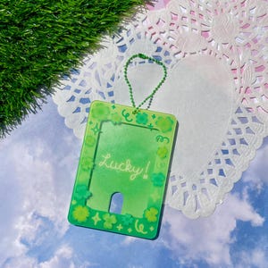 May include: A green card holder with a decorative border of shamrocks and stars. The word "Lucky!" is written in white script across the front. A green beaded chain is attached to the top. The card holder is on a white doily with a blue sky background.