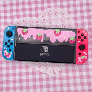 May include: A clear plastic Nintendo Switch case with a pink and white strawberry and dripping icing design. The case has a black Nintendo Switch logo and the words "Nintendo Switch" printed on it.