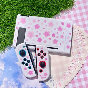 May include: A white video game console dock and two matching controllers featuring a pink cherry blossom pattern. The controllers include black joysticks, directional buttons, and action buttons labeled X, Y, A, and B, with red and blue accent rings.